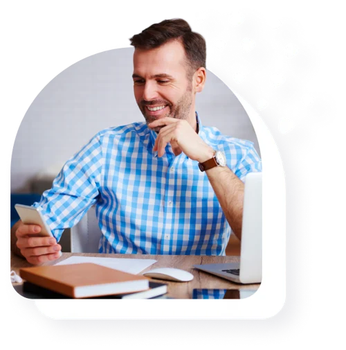 Smiling man in plaid shirt looking at his phone at a desk.